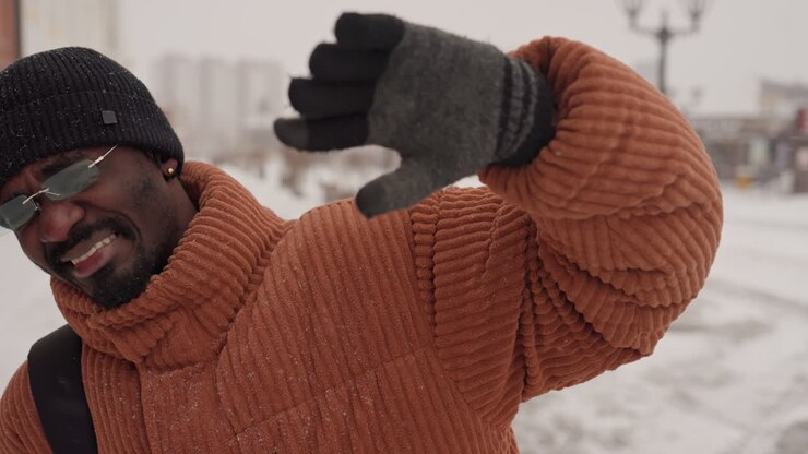 Uomo di colore cammina in una strada innevata, indossa un maglione marrone spesso, un berretto nero, occhiali da sole a specchio e guanti di lana, porta uno zaino mentre si ripara gli occhi dai fiocchi che cadono, vetrine e lampione.