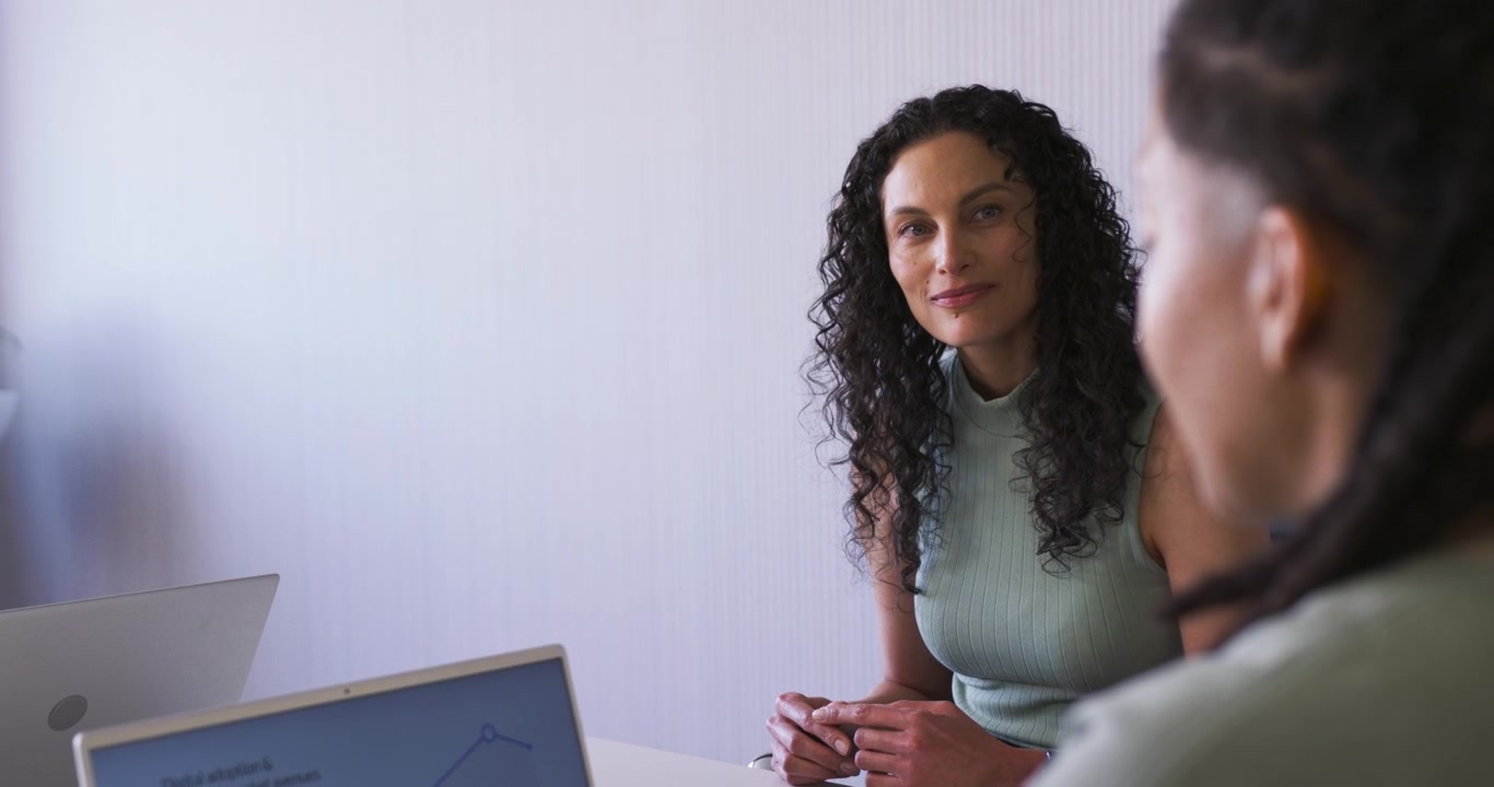 Team afroamericano al tavolo, donna in ascolto mentre una collega con le trecce parla e digita su un grafico al computer