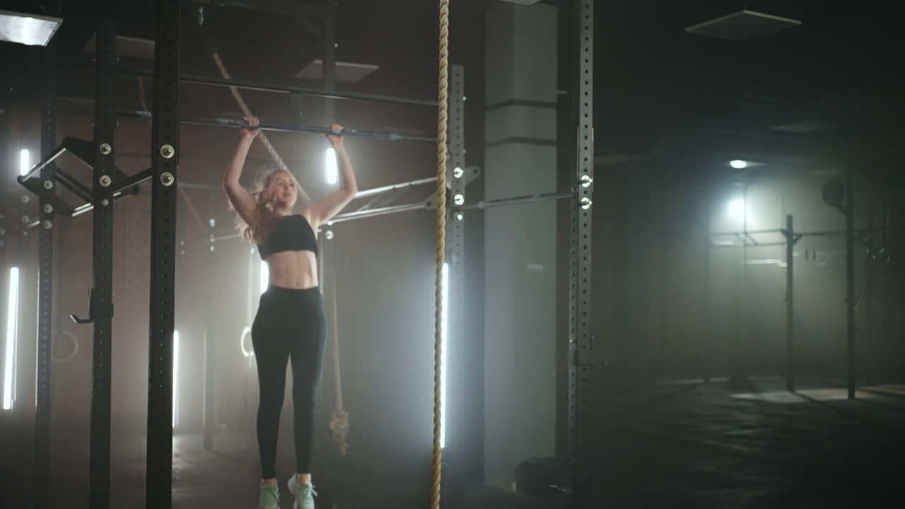 A sporty young woman pulls herself up on a horizontal bar in a dark gym in a beautiful neon backlight. Endurance and perseverance in pulling up movement towards the goal