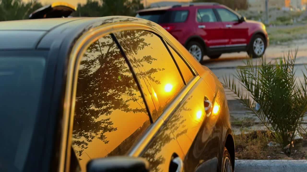 Filmisk nærbillede af en solnedgang i den gyldne time, der reflekteres i en sort bils ruder på en udendørs parkeringsplads. Levende orange lys gløder mod den slanke køretøjsoverflade i skumringen.