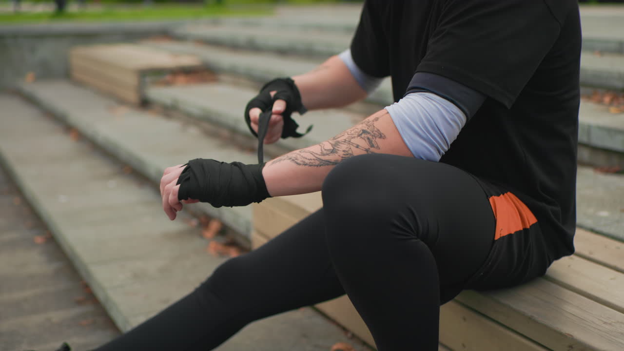 Junger, weißer Mann bandagiert seine Handgelenke auf Außentreppen, sitzend auf Betonstufen, schwarzes Shirt mit orangenen Shorts und Leggings, sichtbare Unterarmtätowierungen, Nahaufnahme der Hände beim Anbringen der Bandagen, ruhig und konzentriert