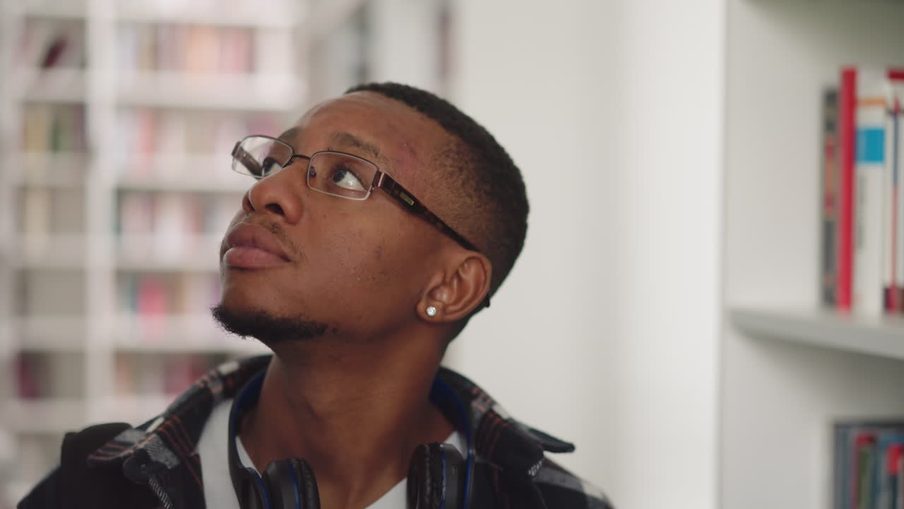 estudiante con gafas mira a su alrededor en la biblioteca de la universidad. hombre afroamericano pensativo busca literatura en la librería. fuentes de educación para el trabajo