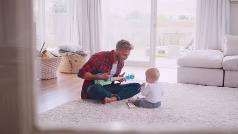 Papá-Sentado-Tocando-El-Ukelele-Con-Un-Niño-Pequeño,-Visto-A-Través-De-La-Ventana