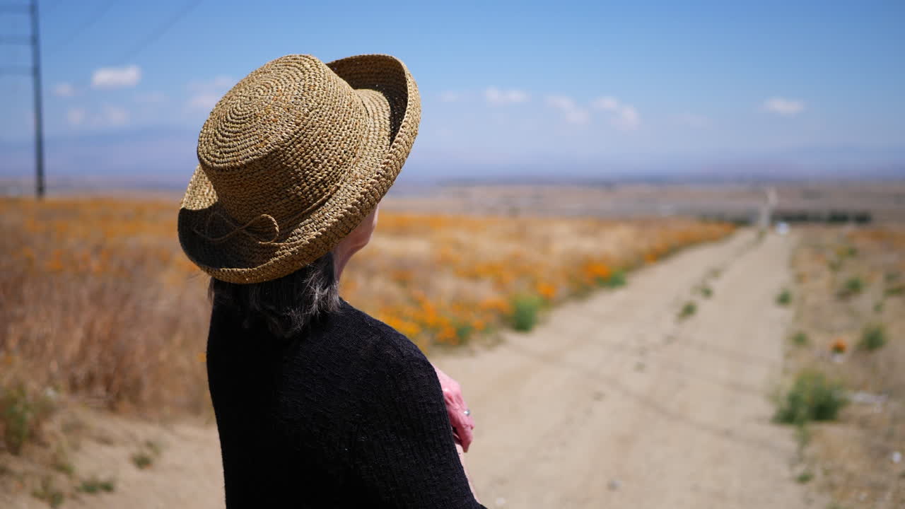 An old woman in a summer hat pointing down a dirt road hitchhiking in a deserted field of orange California poppy flowers SLOW MOTION