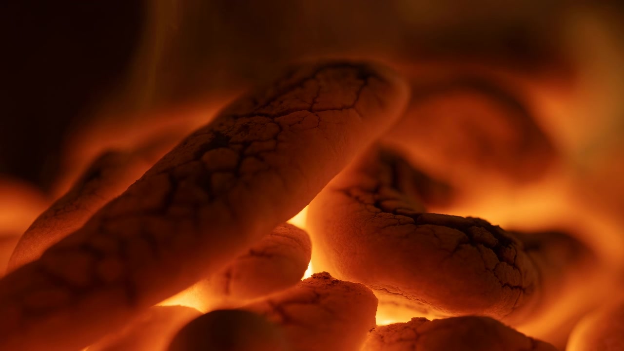 Vista ravvicinata dal basso di braci di carbone impilate che mostrano un ardere attivo, un profondo bagliore interno, una fiamma debole e una consistenza cenere friabile in un calore rustico scuro con forte contrasto e profondità stratificata.