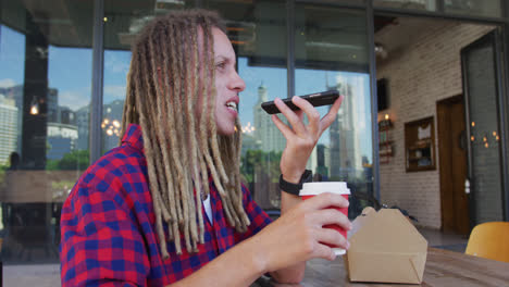 mixed race man with dreadlocks sitting at table outside cafe drinking coffee and using smartphone