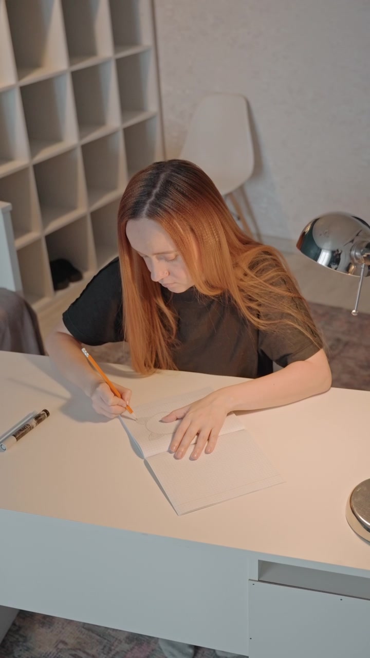 Young woman with reddish brown hair is seated at white desk. Woman uses orange pencil and occasionally steadies sheet. Scene features consistent lighting from natural sources