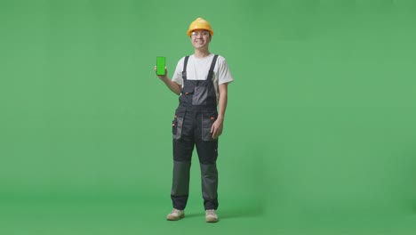 cuerpo lleno de hombre asiático trabajador con gafas de protección y casco de seguridad sonriendo y mostrando pantalla verde teléfono inteligente a la cámara mientras estaba de pie en la pantalla verde estudio de fondo