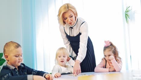 three little children with the teacher in class. beautiful young woman teaching english at school. teacher helps elementary students with pictures.