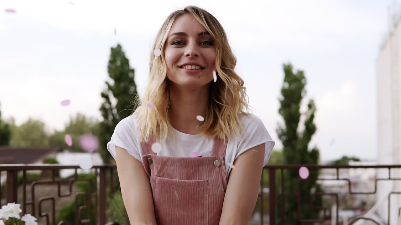 A cute, attractive woman with natural beauty blowing confetti in slow motion. Pink and blue colored. Blonde haired caucasian. Blurred trees on the background. Outdoors
