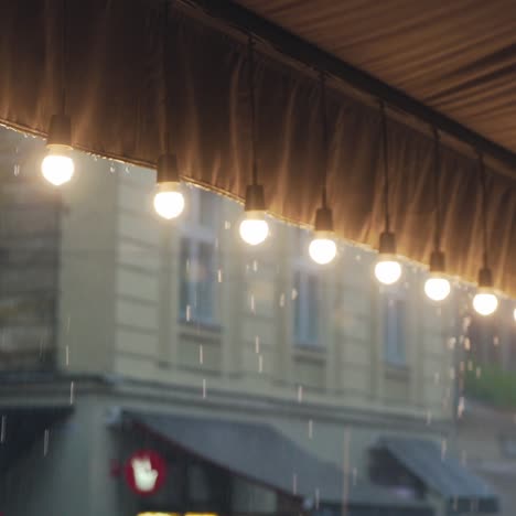 decorative lamps are hanging in the row and lighting on the background of the rain. raindrops are falling on the roof of the summer terrace of a cafe which is decorated with a row of lanterns