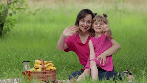 family weekend at picnic. daughter child girl with mother sit on grass meadow and waving hands