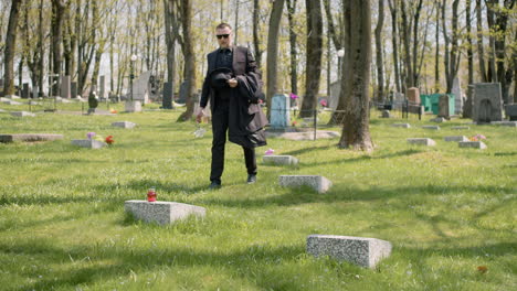 Trauriger-Mann-Im-Schwarzen-Anzug,-Der-Auf-Einem-Friedhof-Spaziert,-Die-Sonnenbrille-Abnimmt-Und-Eine-Weiße-Blume-Auf-Einen-Grabstein-Legt-1