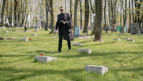 hombre triste con traje negro caminando en un cementerio, quitándose las gafas de sol y colocando una flor blanca en una lápida 1