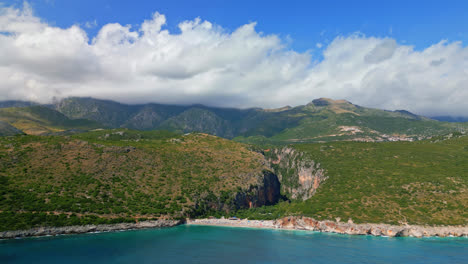 aerial drone shot flying high in distance over gjipe beach, albania with mountain range in the background on a sunny day