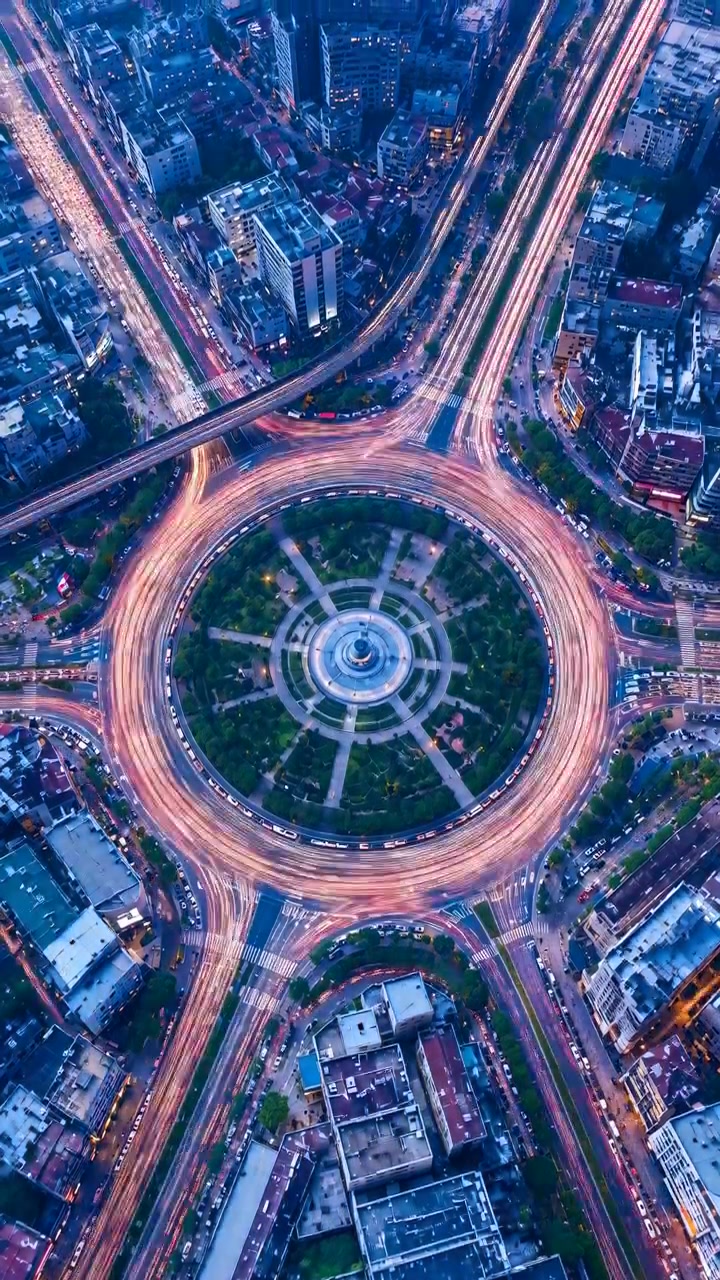 Centered night aerial shows large circular roundabout with red and white light trails circling landscaped center island, illuminated fountain, layered overpasses, and glowing urban roads
