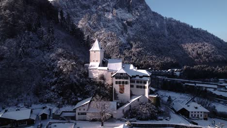 Luftaufnahme-Eines-Bergdorfes-Mit-Einer-Kirche-Und-Schneebedeckten-Felsigen-Bergen-Im-Hintergrund-In-Der-Schweiz-An-Einem-Sonnigen-Wintertag