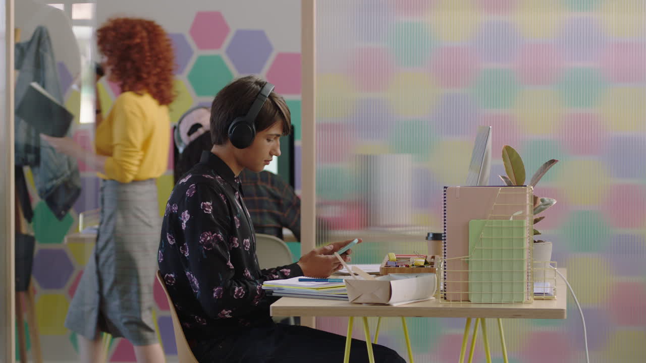 젊은 백인 남성 학생이 스마트 폰을 사용하여 문자 메시지 소셜 미디어 브레인 스토밍 다채로운 현대 사무실에서 창의적인 프로젝트를 진행하고 헤드폰을 착용하여 음악을 듣고 있습니다.