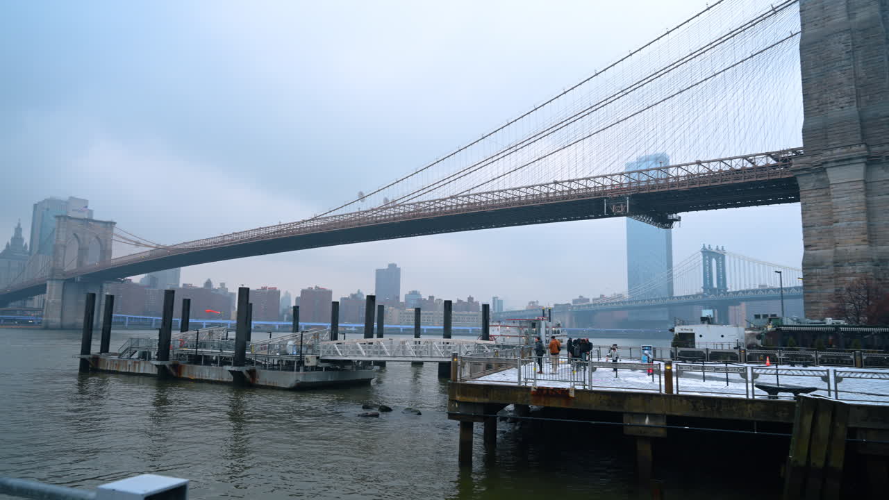 New York, USA, 28 dicembre 2025: Persone che passeggiano in inverno vicino al ponte di Brooklyn e all'East River a NYC. La gente si raduna vicino all'acqua nei pressi del ponte di Brooklyn in una giornata invernale nuvolosa a New York
