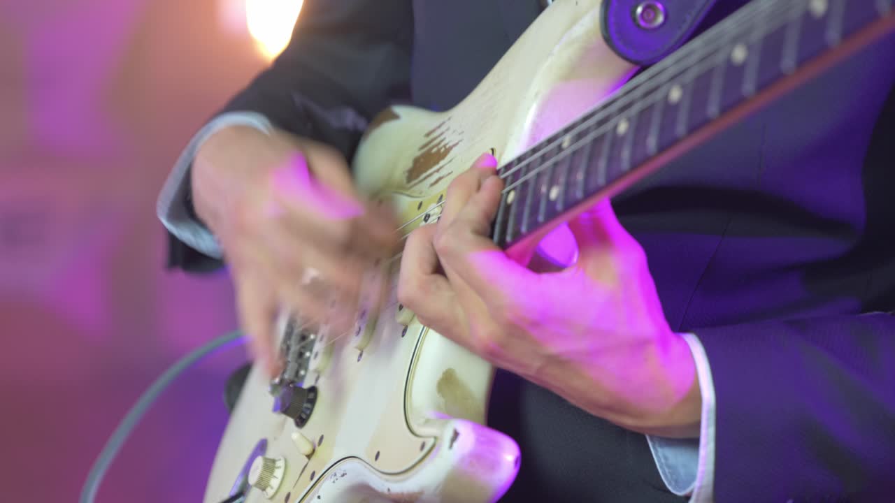 Primo piano di un musicista professionista che suona accordi veloci su una chitarra elettrica Stratocaster durante una sessione dal vivo sul palco, con luci calde da studio sullo sfondo sfocato.