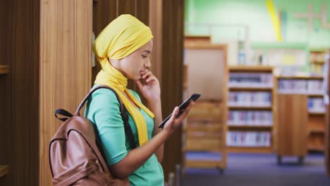 Asian-female-student-wearing-a-yellow-hijab-leaning-against-bookshelves-and-using-tablet