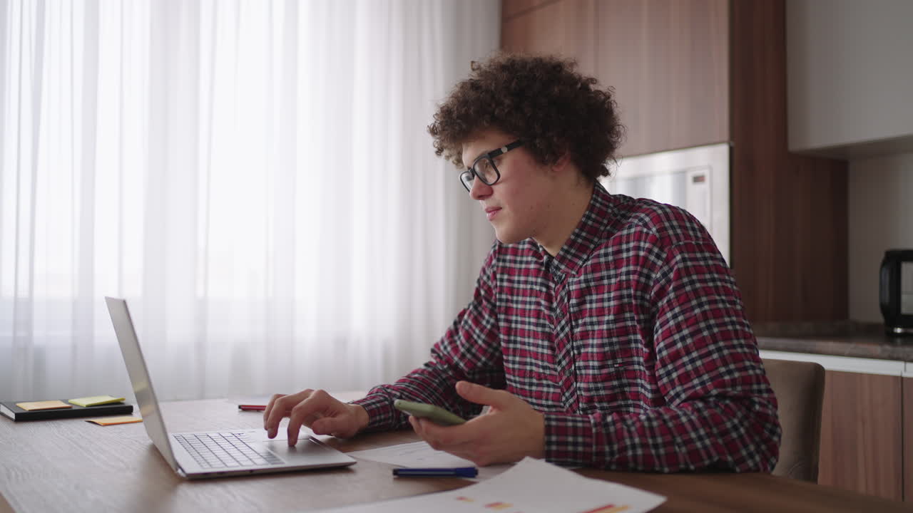 Curly haired Male student attractive young boy in glasses is studying at home using laptop typing writing in notebook. College student using laptop computer watching distance online learning seminar