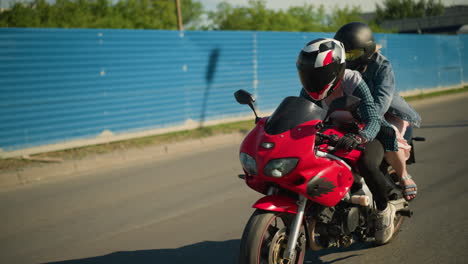 two friends ride a red power bike wearing helmets, passing a construction site with blue zinc sheets, cars with headlights on are seen approaching in the background near a city building