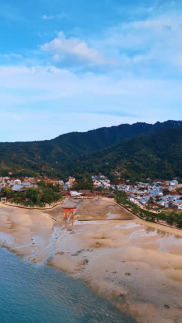 flight above the waterscape approaching a beautiful red gates on the shore with multiple tourists walk around. aerial perspective on the stunning itsukushima shrine, japan. vertical video.