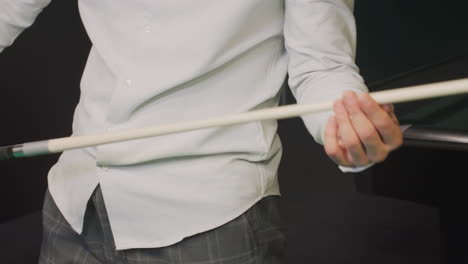 Male in white shirt and grey trousers, face not visible, picking up cue stick beside green pool table. Focus on hands, billiard balls, precision grip, professional setup, competitive atmosphere