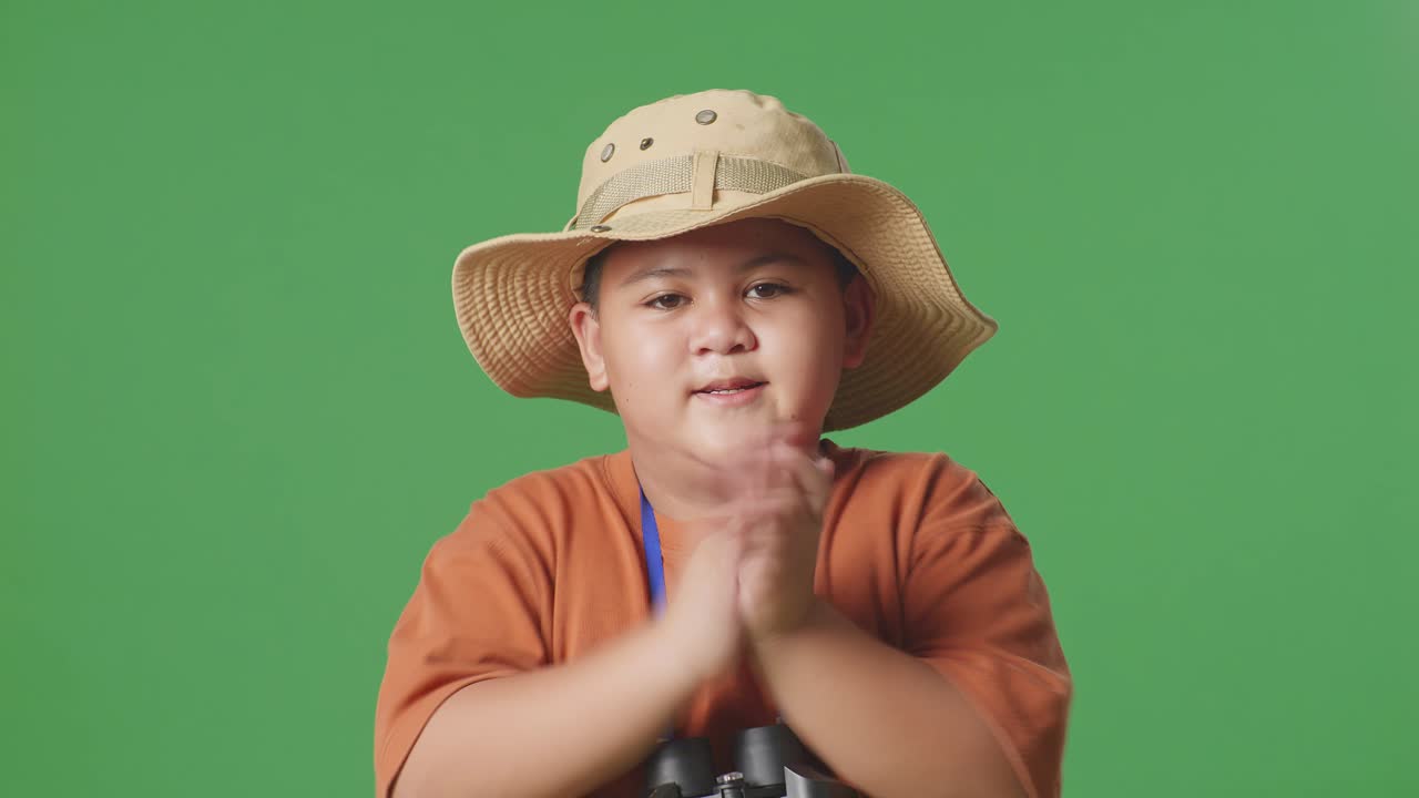 niño turista asiático con un sombrero aplaudiendo las manos después de mirar a través de los binoculares. niño investigador examina algo en el fondo de la pantalla verde, concepto de aventura de turismo de viaje, de cerca