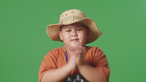 모자를 입은 아시아 관광 소년은 안경을 통해 쳐다본 후 손을 치고 있습니다. 소년 연구자는 녹색 화면 배경에서 무언가를 조사합니다. 여행 관광 모험 개념, 근접