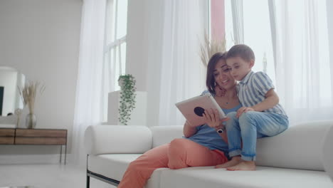 madre e hijo sentados en el sofá usando tableta digital. madre feliz y niño pequeño usando tableta con pantalla táctil juntos viendo un video. madre sonriente y niño lindo jugando en tablet digital.