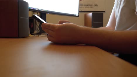 guy receives and checks a snapchat push notification on his cellphone while sitting at his home office desktop computer