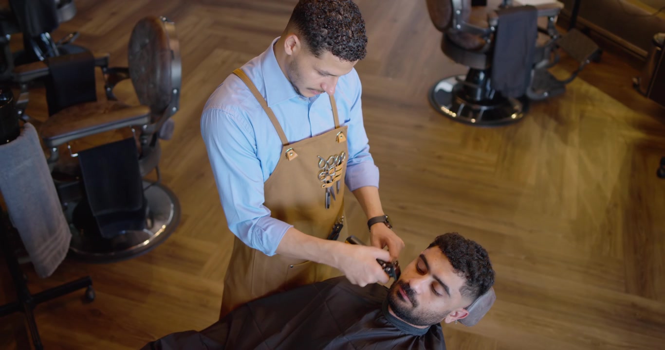 Mandlig barber trimmer en tilbagelænet mandlig kundes ansigtshår med en trimmer for at forme skægget i en barbersalon.