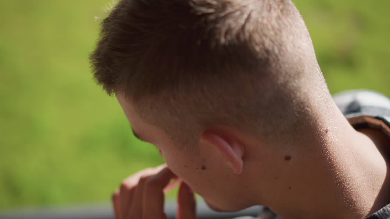 rear view of young man in checkered shirt on outdoor staircase retrieving cigarette from ear tilts head forward and raises lighter to lips against softly blurred greenery background