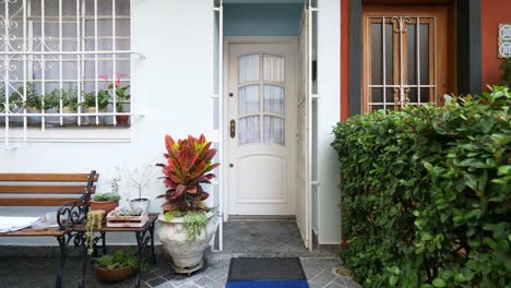 a white metal gate with ornate patterns is open at a house entrance. nearby, umbrellas and rain boots rest on a tiled bench under a matching window grill and wall lamp.