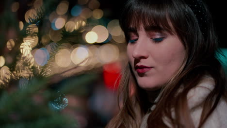 primer plano cinematográfico de una hermosa mujer mirando un árbol de navidad y sonriendo rodeada de luces de navidad en un mercado de navidad al fondo