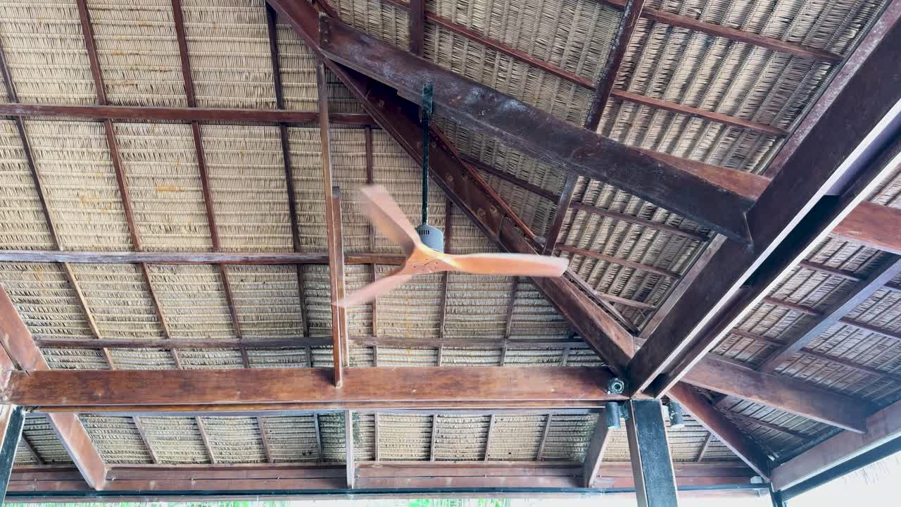 A wooden ceiling fan rotates under a rustic, open-air roof in Phuket, Thailand. Natural lighting highlights the wooden beams and fan blades