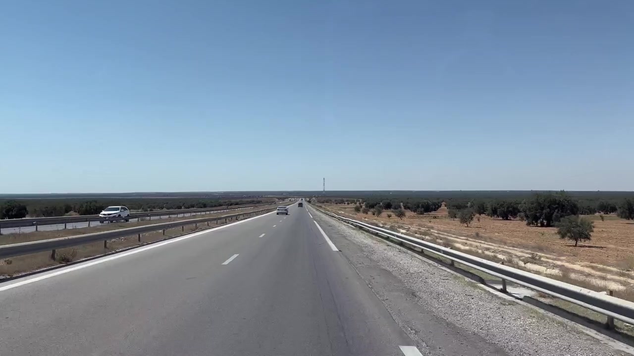 Auto viaggiano lungo un'autostrada a carreggiate separate che attraversa terreni agricoli piatti e aridi sotto un cielo azzurro e limpido, con guardrail che incorniciano la strada vuota davanti in Tunisia.