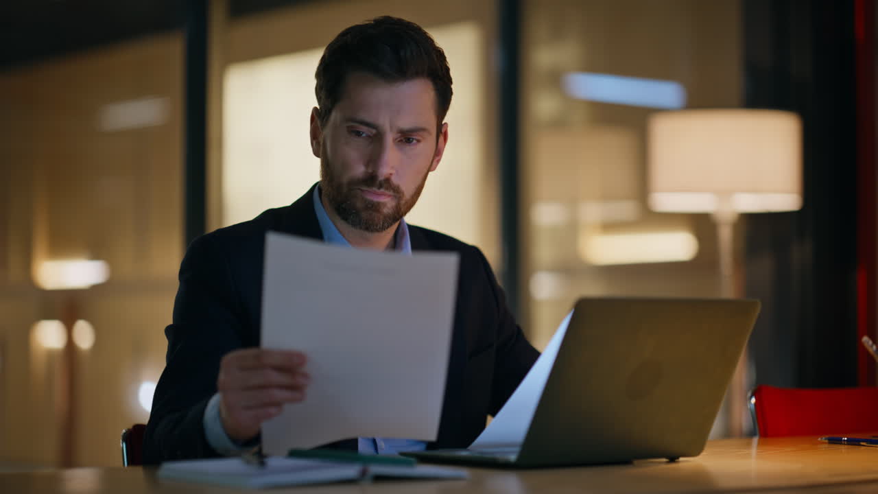 Late worker reading papers working at laptop in dark office closeup. Serious man