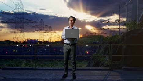 full body of an asian male professional worker standing with his laptop near high voltage tower, industrial facility, he observes by looking around before he come to concentrating with his laptop