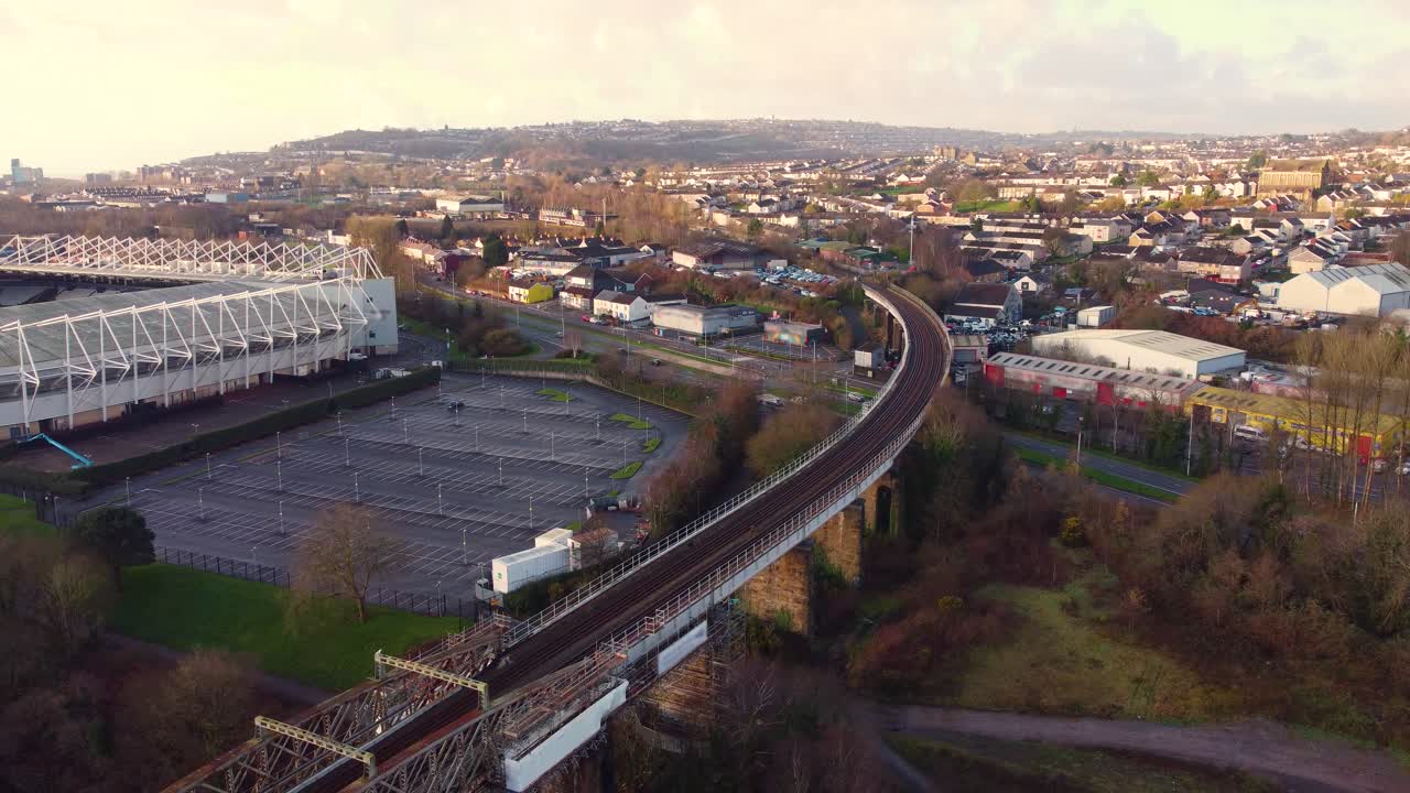 Langsomt sideværts luftfoto med snoet togbro i Swansea og omkringliggende huse med Swansea Football Stadium. 21.000-sæders skålformet arkitektur, omkringliggende parkeringspladser.