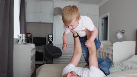 dad keeps a dignity above himself lying on the bed. a boy in a white t-shirt laughs and smiles from playing with his father