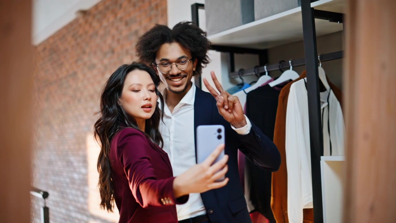 Foto selfie de una pareja sonriendo en una tienda de ropa. Mujer con teléfono y hombre haciendo señal de paz en la tienda
