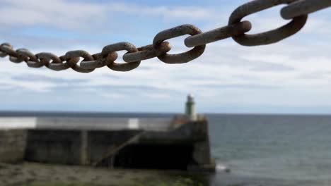 chain link in focus with waves hitting stone pier with small lighthouse at laxey, isle of man