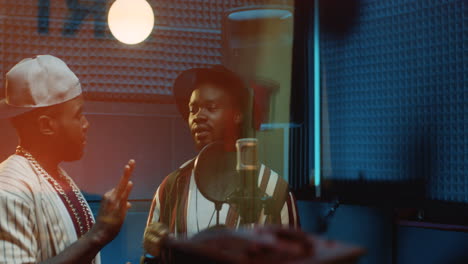 Two-Young-Stylish-Male-Singers-Or-Rappers-Standing-At-The-Microphone-In-The-Dark-Sound-Studio-Over-The-Glass-Window-And-Chatting-While-Recording-A-Common-Hit-Song