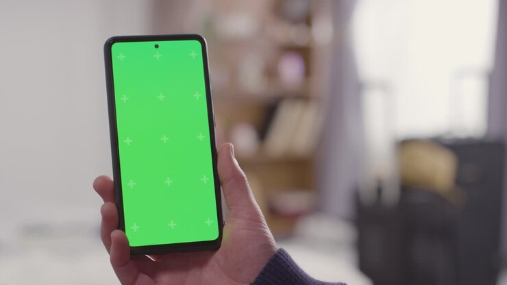 Persona a casa che tiene in mano un telefono cellulare con schermo verde, con valigia preparata per le vacanze estive sullo sfondo