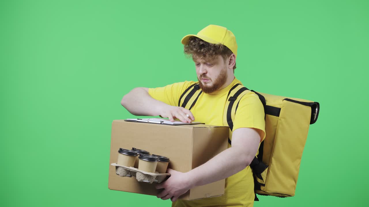 Portrait of young men fills out delivery documents holding parcel and coffee. Redhead male in yellow courier uniform with thermo bag posing on green screen. Close up. Slow motion ready 59.97fps