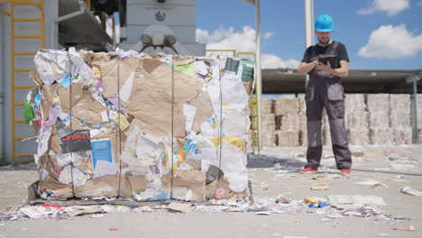 Man-in-hardhat-uses-tablet-by-paper-bale-at-recycling-plant,-static