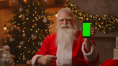 kind santa claus is holding a smartphone with a green screen. indicates the smartphone screen. sits in a beautiful room decorated for a merry christmas.
